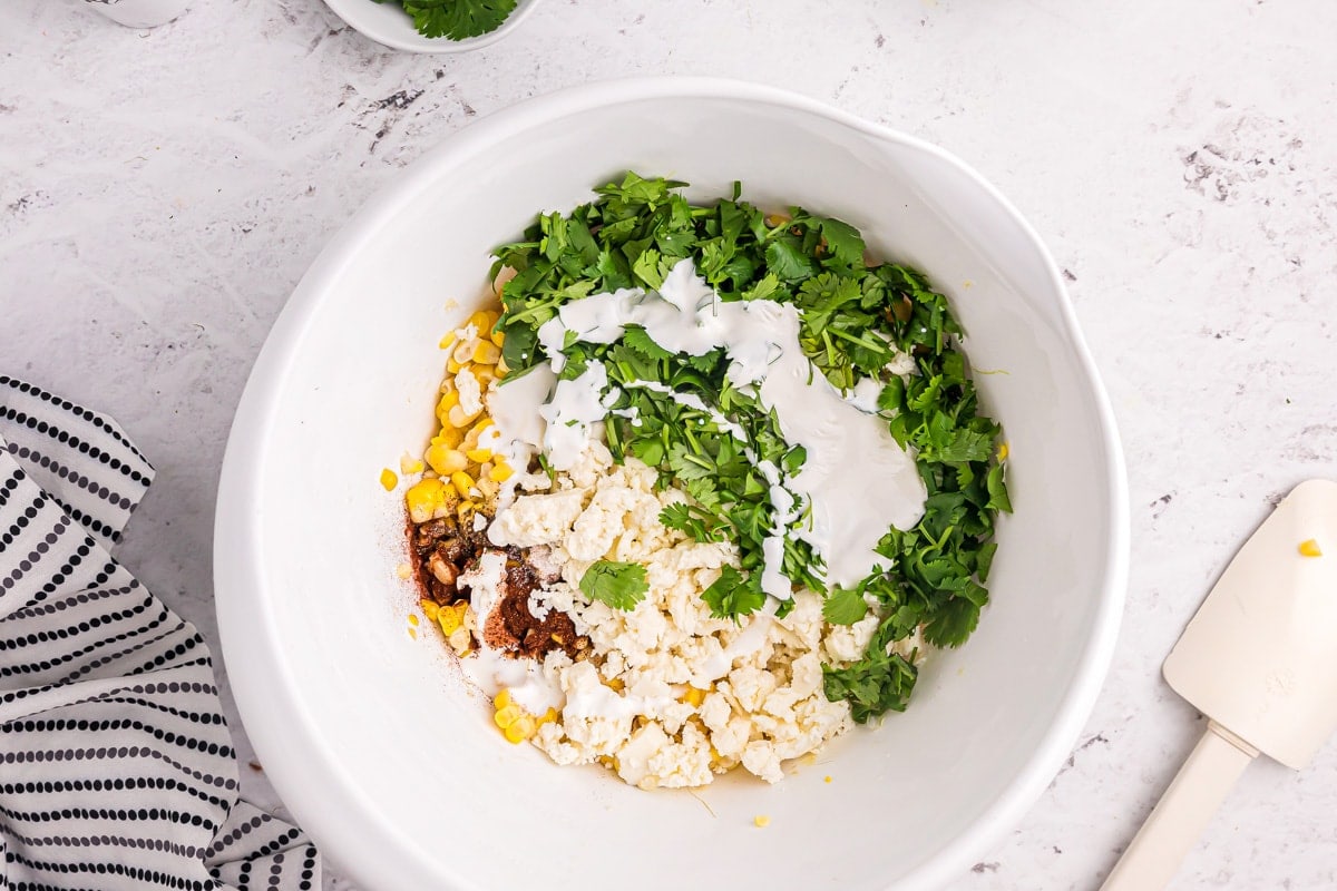 Combining corn, cheese, herbs, and seasonings in a white bowl on a kitchen counter.