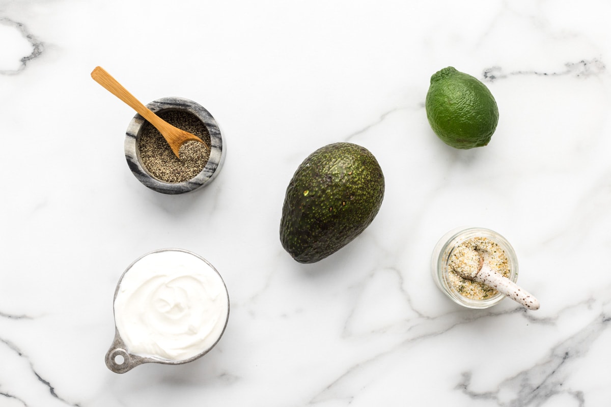 Avocado, lime, sour cream, and seasonings on a kitchen counter.