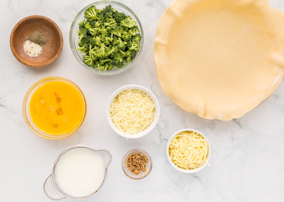 A pie crust, measured cheese and broccoli, and eggs measured on a kitchen counter.