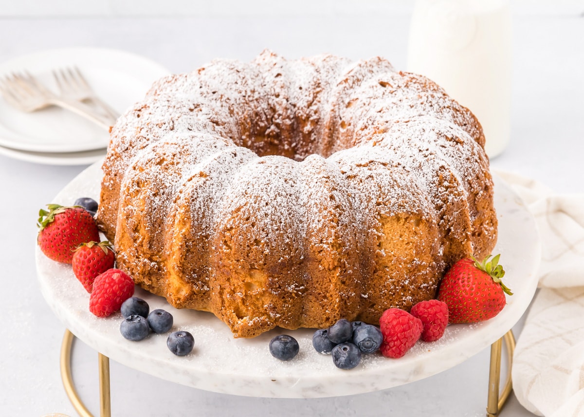 A cream cheese pound cake recipe on a cake stand surrounded by fresh fruit.