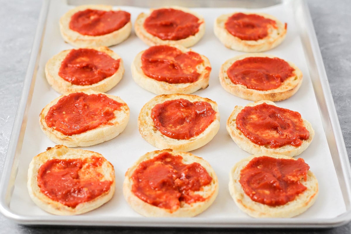 Adding sauce to toasted english muffins on a lined baking sheet.