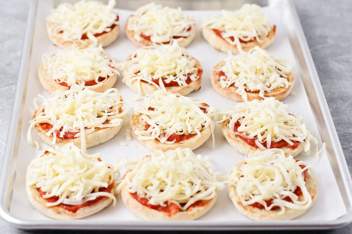 Adding cheese to english muffins on a lined baking sheet.