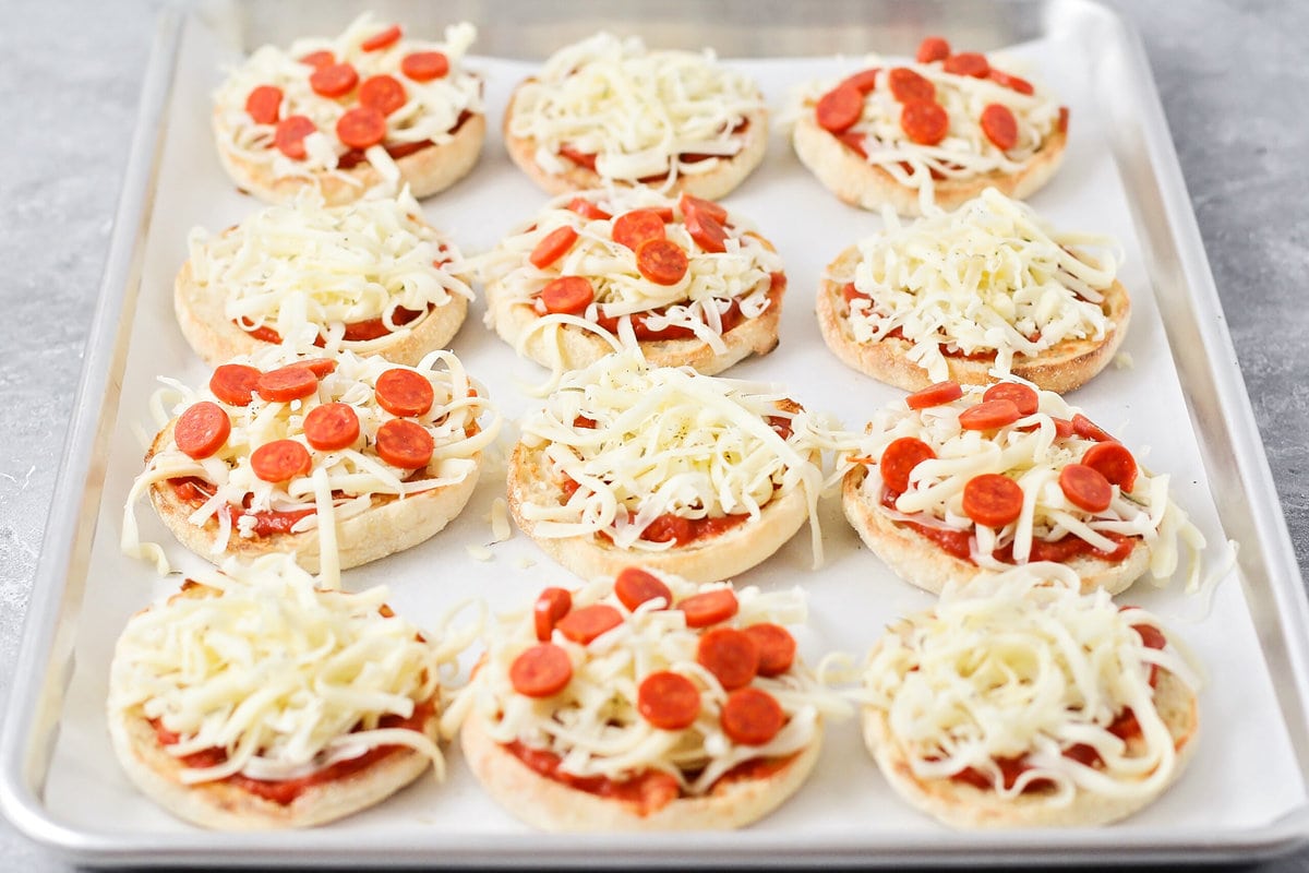 Adding pepperoni to english muffins on a lined baking sheet.