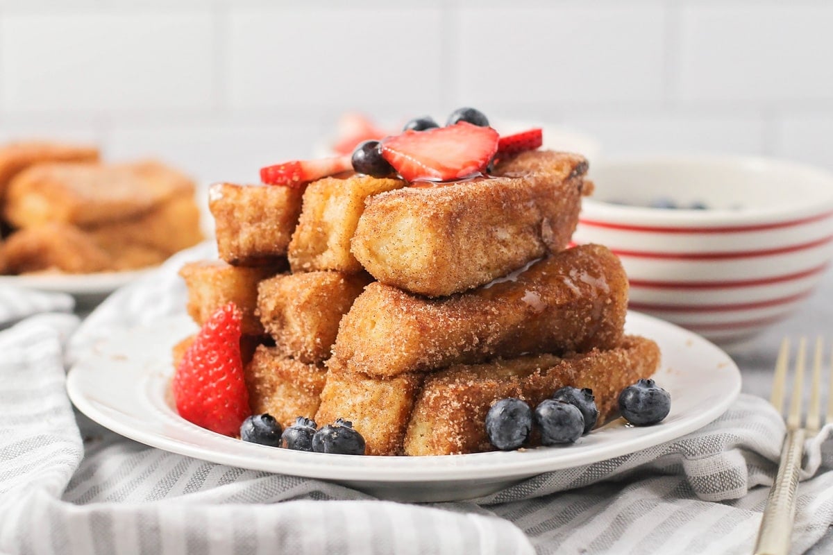 Several french toast sticks stacked on a white plate with fresh fruit.