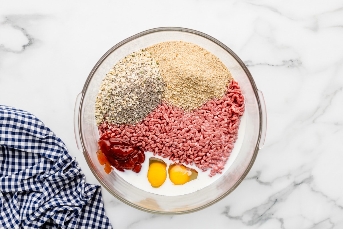 Adding eggs, milk, and seasonings to ground beef in a glass bowl.