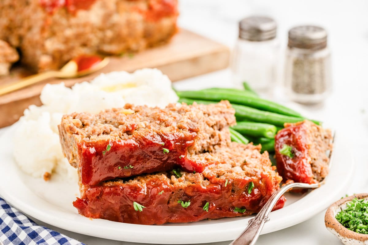 Two slices of meatloaf on a plate with mashed potatoes and green beans.