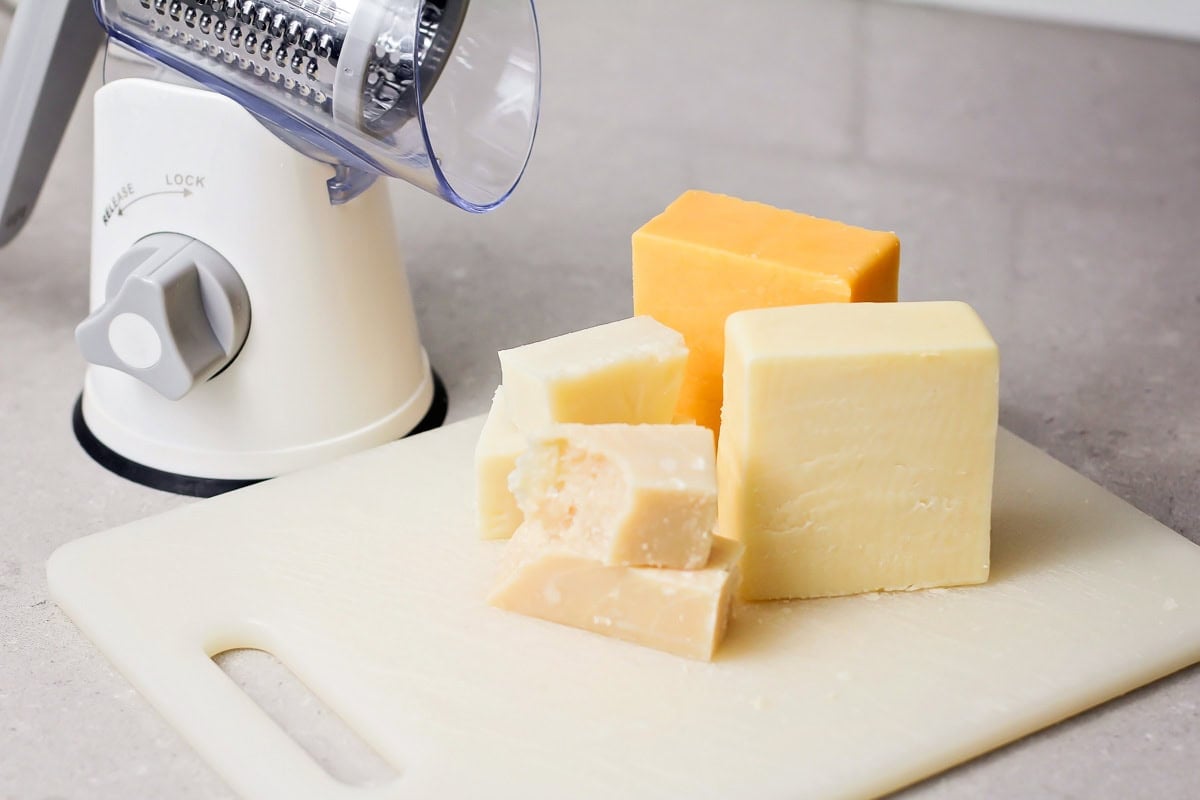 Three different types of cheese on a cutting board with a grater.