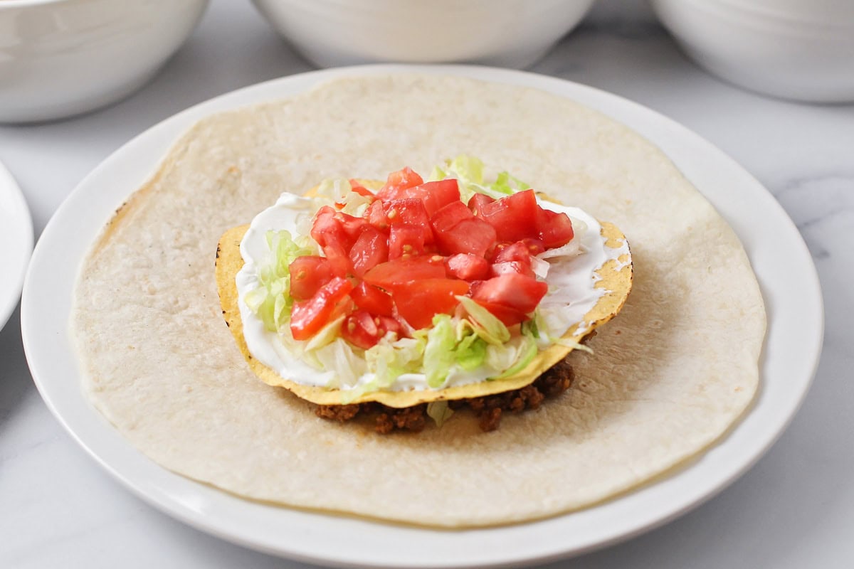 Layering meat, nacho cheese, tostada, sour cream, and veggies on a large tortilla.