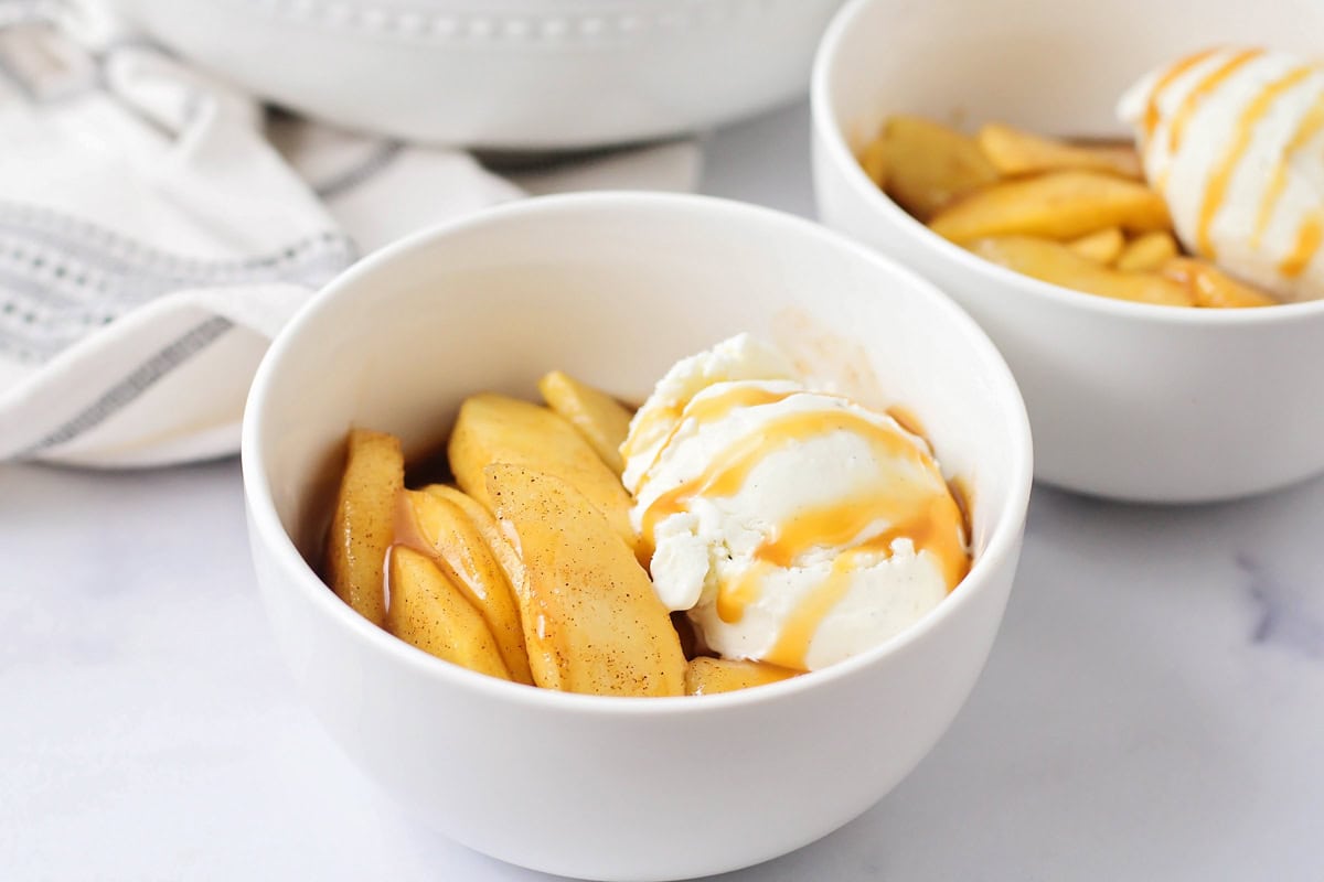 Fried apples served in a white bowl with  a scoop of vanilla ice cream and caramel sauce.