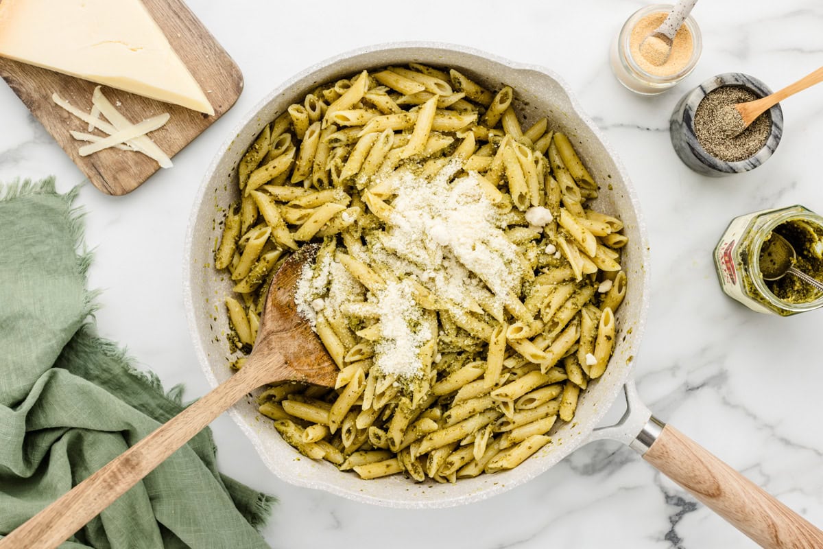 Adding parmesan to pesto and cooked pasta in a skillet.