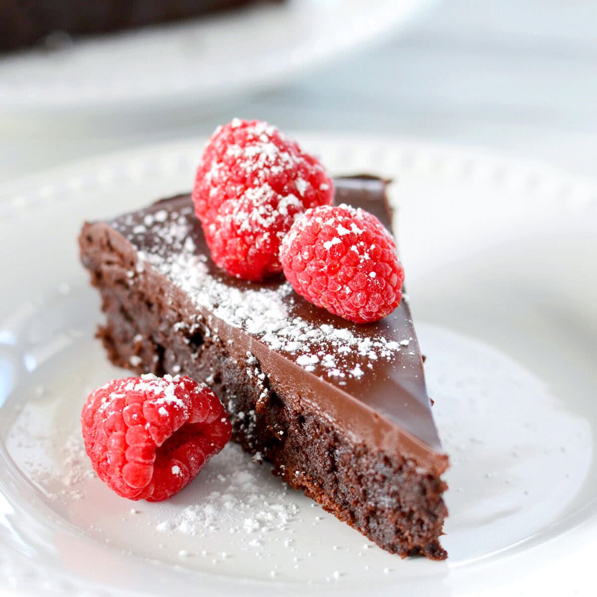 Slice of flourless chocolate cake sprinkled with powdered sugar and topped with fresh raspberries.