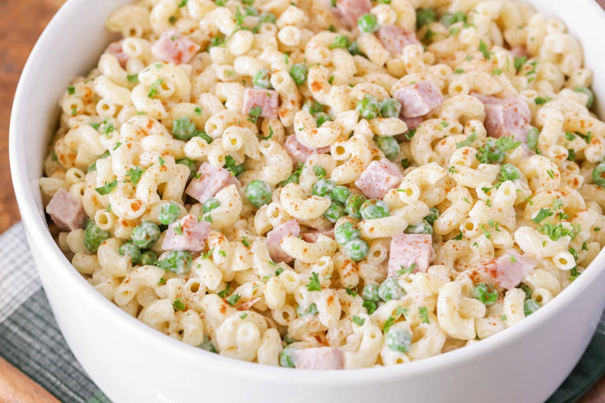 Close up image of macaroni salad in white bowl with peas and ham cubes.