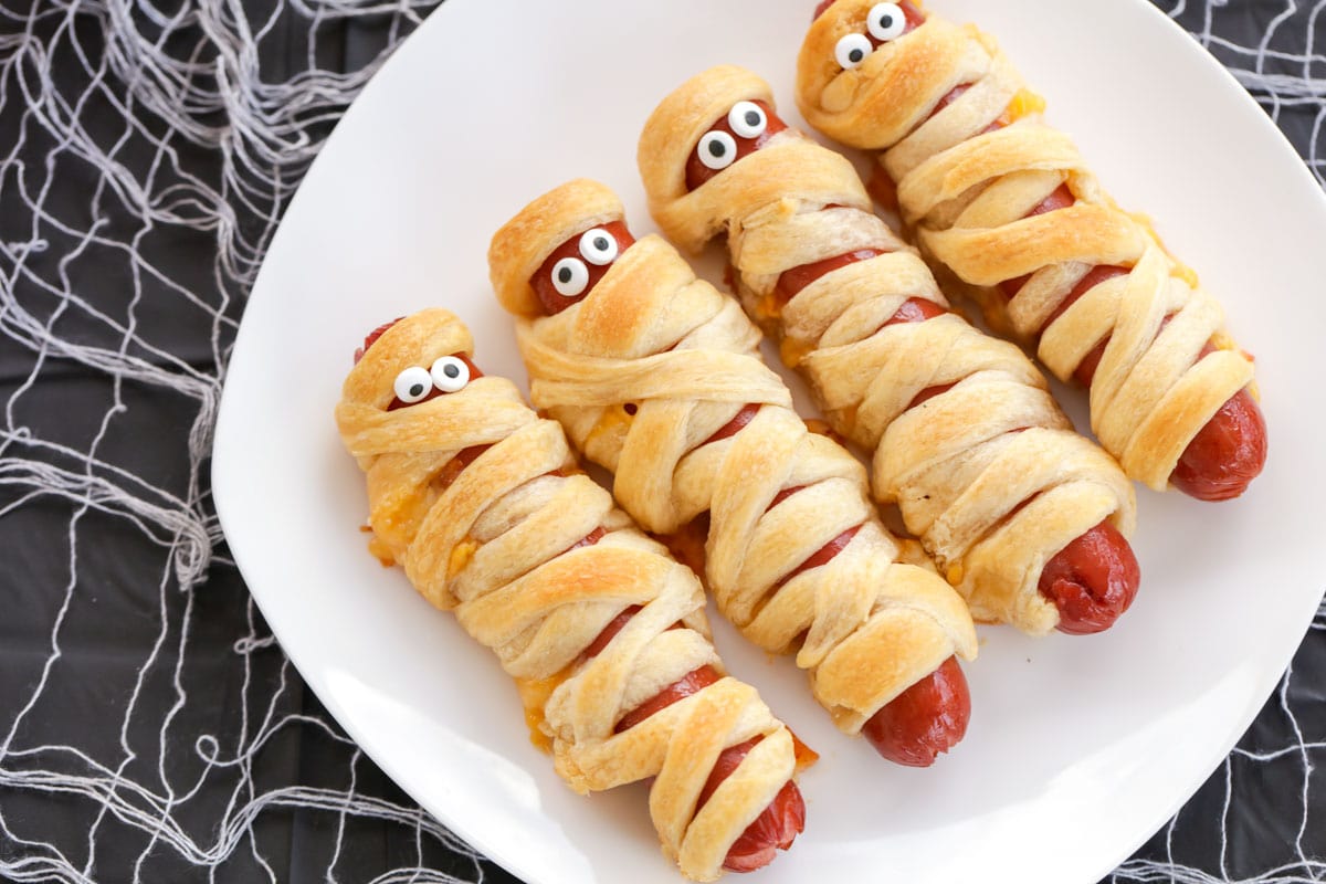 4 Mummy Hot dogs on white plate on black tablecloth.