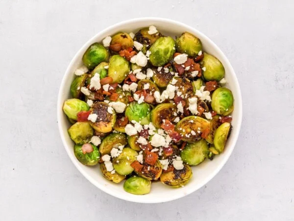 Thanksgiving Brussel sprouts sprinkled with bacon and goat cheese in a white bowl.