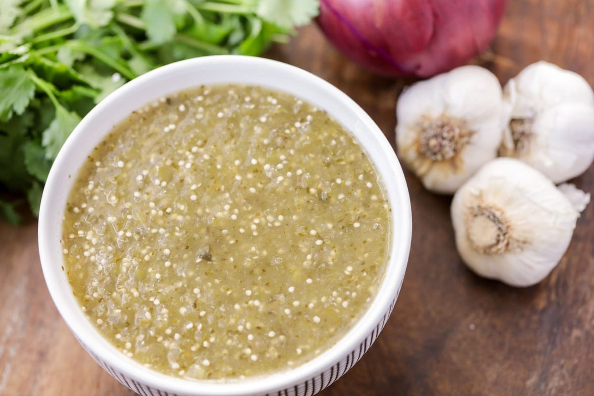 A bowl of tomatillo salsa with whole garlic bulbs, red onion, and cilantro next to it. 