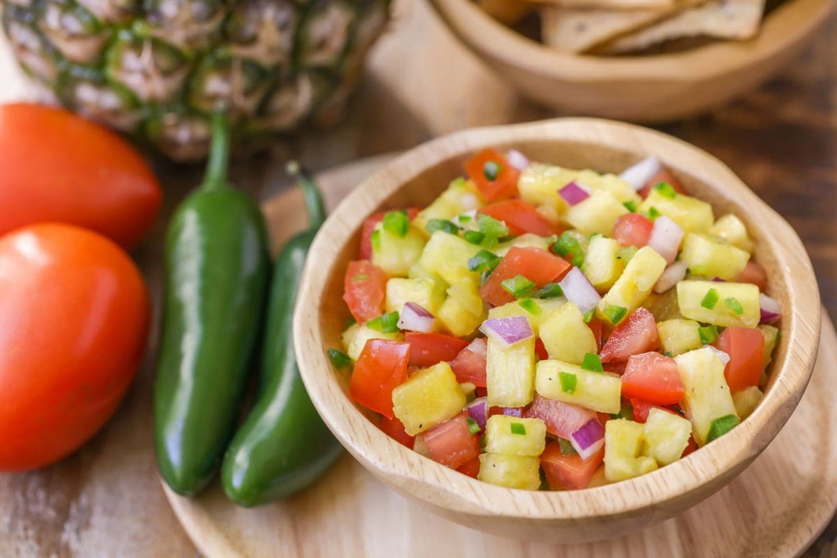 a bowl of pineapple salsa with fresh jalapenos, tomatoes, and a pineapple next to it.