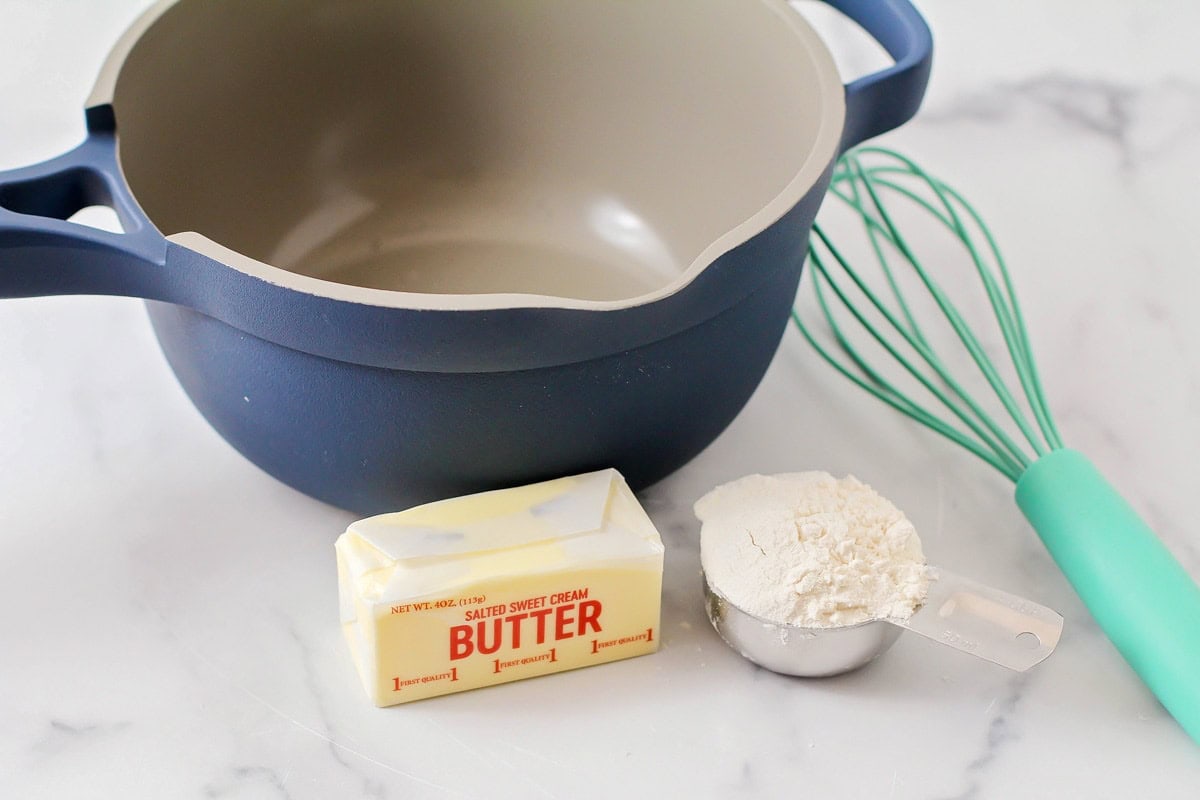 A navy blue pot with a stick of butter, measuring cup flour, and a light blue whisk sitting next to it. 