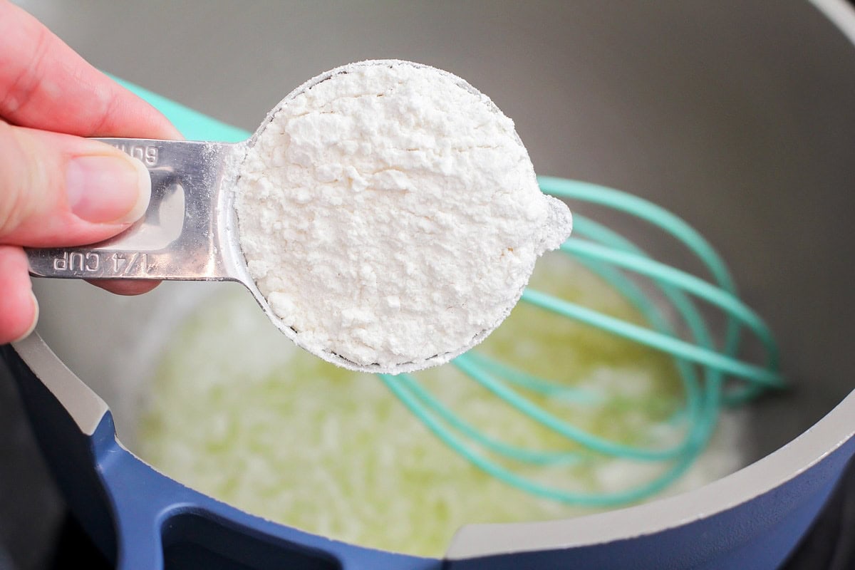 A measuring cup of flour pictured with melted butter in a pot and a whisk in the background. 