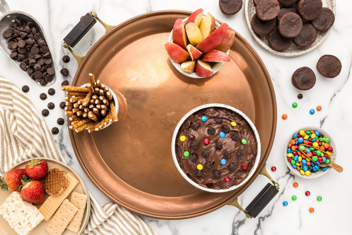 Adding containers of brownie dip, pretzels, and apples to a round tray.