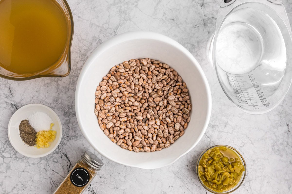 A bowl of dried beans with a glass measuring cup full of broth, a small bowl of spices, and a jar of cumin beside it. 