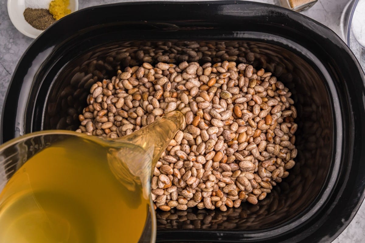 A slow cooker full of dried beans with broth being poured into it. 