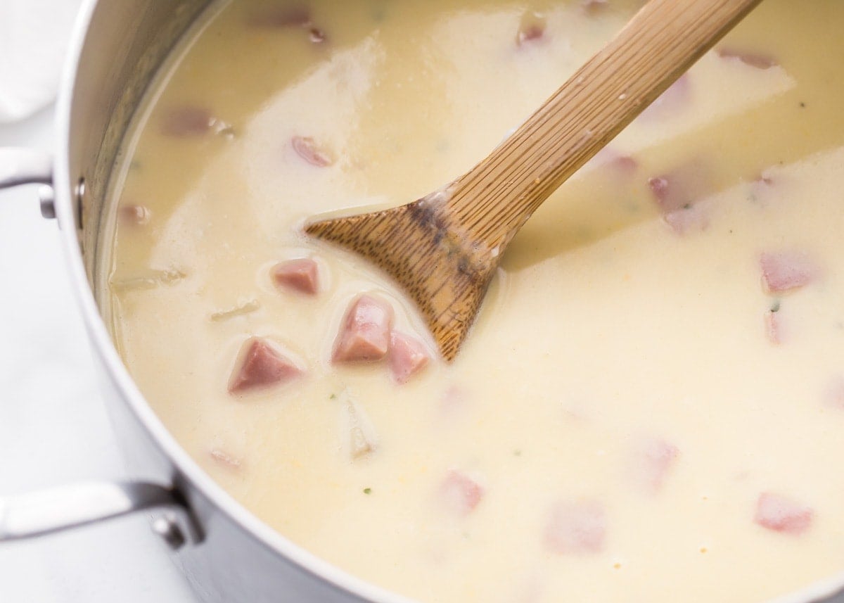 A close up photo of a pot of ham and potato soup with a wooden spoon stirring the soup. 