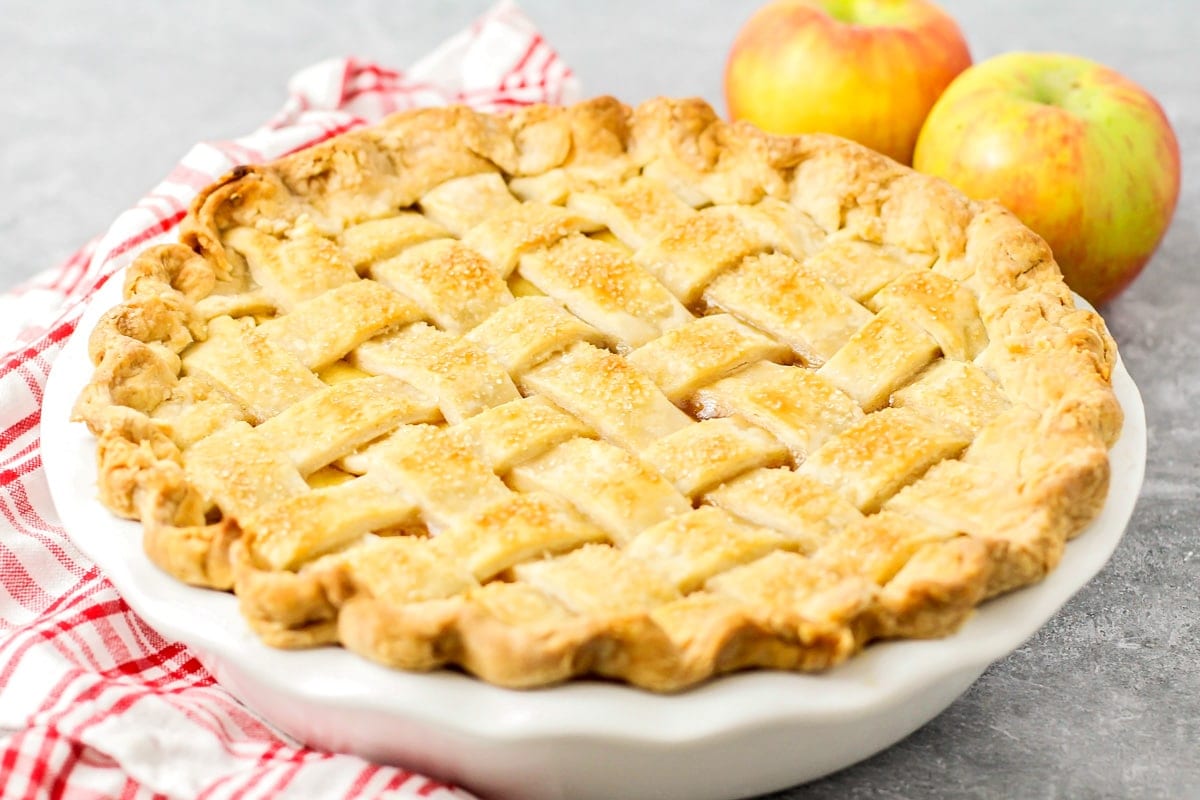 A baked apple pie with a lattice pie crust alongside of two fresh apples. 