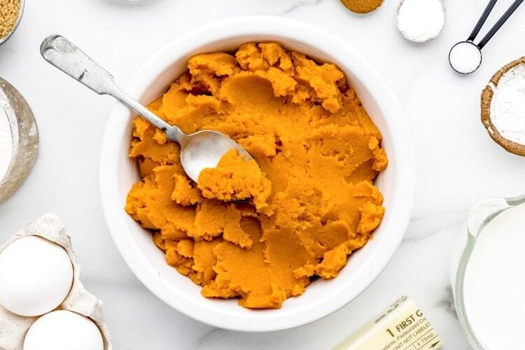 A bowl of pumpkin puree surrounded by other ingredients in measuring spoons and measuring cups, eggs, and a stick of butter.