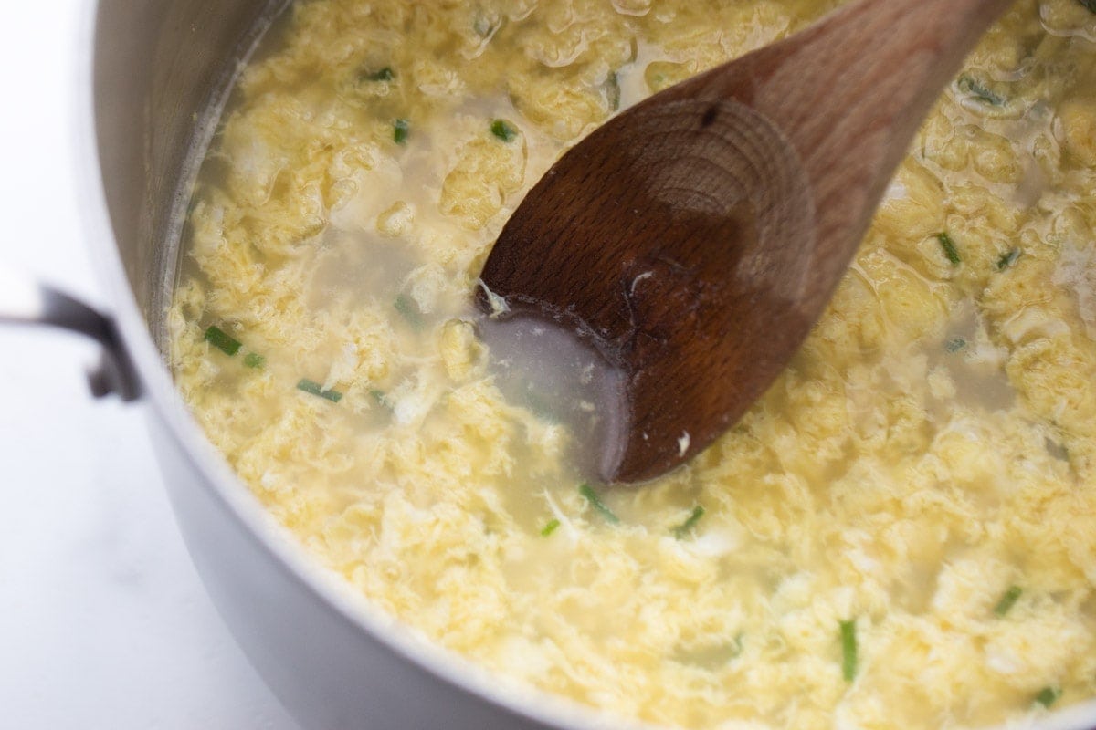 A close up photo of egg drop soup in a pot being stirred with a wooden spon