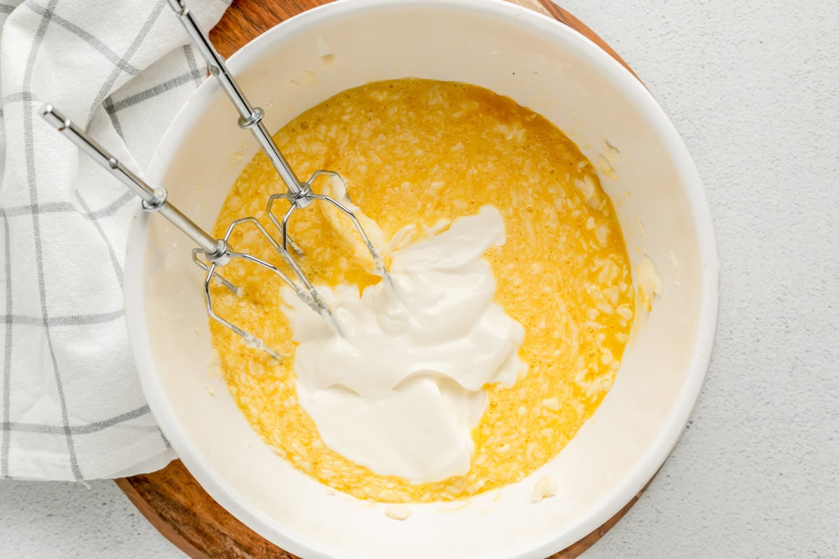 Sour cream being mixed into corn pudding mixture in white bowl.