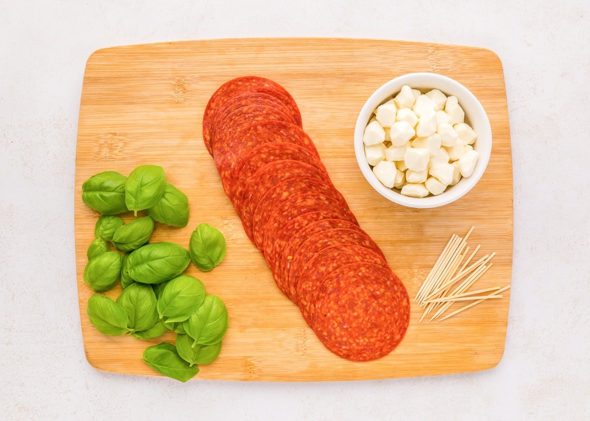 Pepperoni, mozzarella pearls, and basil leaves on a wooden cutting board.