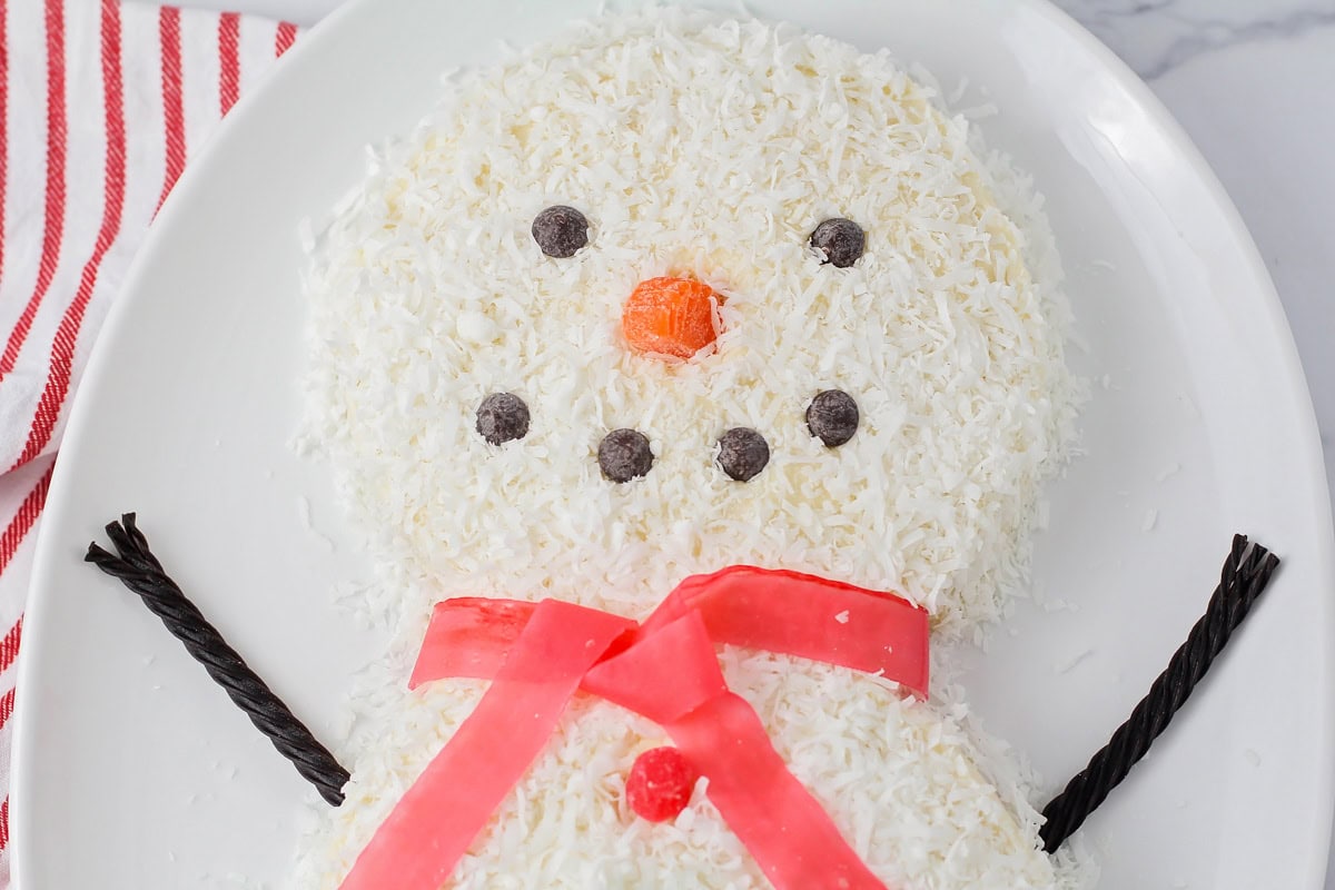 A snowman cake with chocolate chip eyes and mouth and candy arms and scarf.