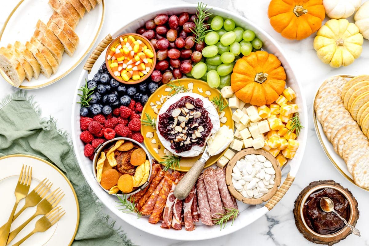 A charcuterie plate with purple and green grapes, cheese cubes, assorted meats, berries, candy corn, decorated with small pumpkins. Crackers and bread are on the side along with jam.