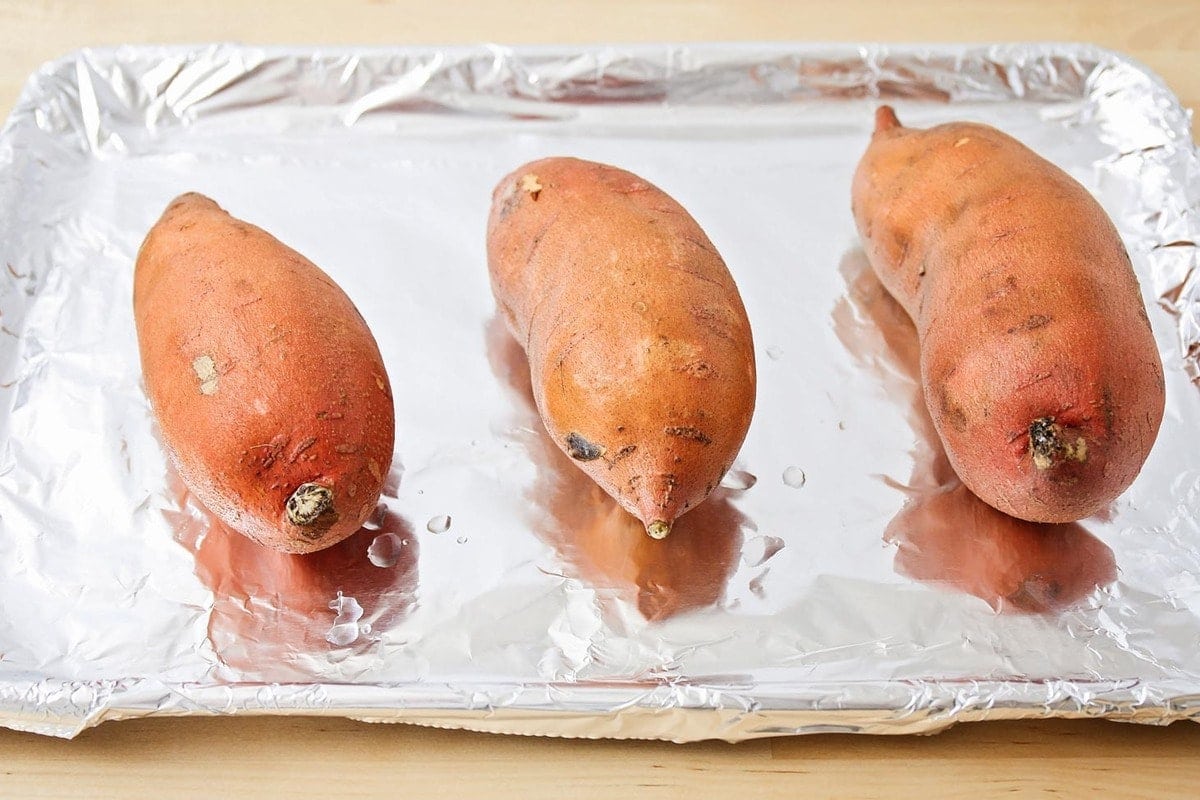 A sheet pan lined with foil with three sweet potatoes on it. 