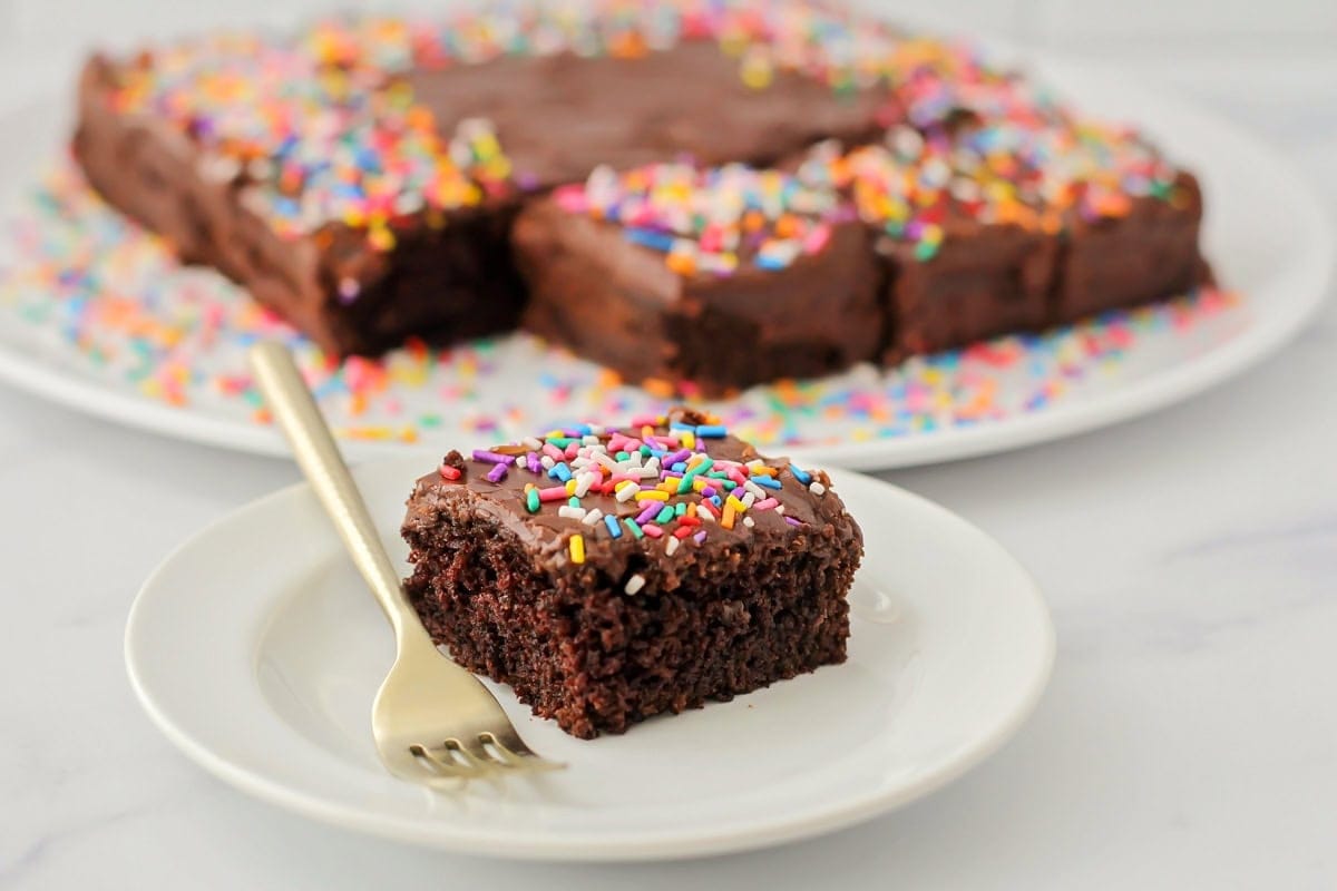 A piece of chocolate cake on a dessert plate topped with rainbow sprinkles with the cake blurred in the background. 