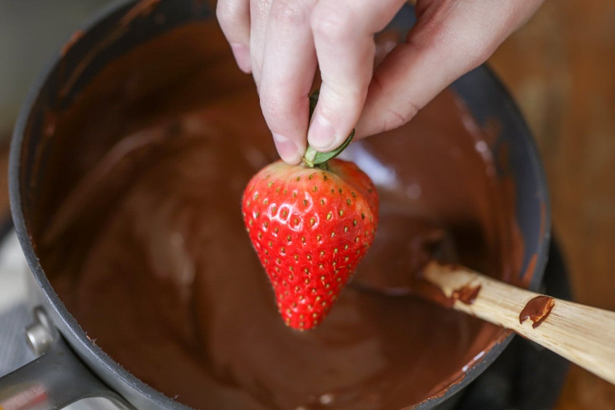 A hand holding a strawberry over a pot of melted chocoalte