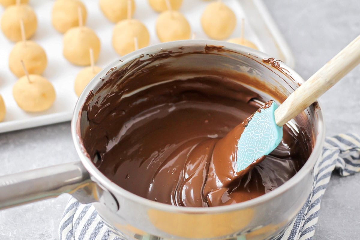 A saucepan of melted chocolate with buckeyes in the background ready to be dipped. 