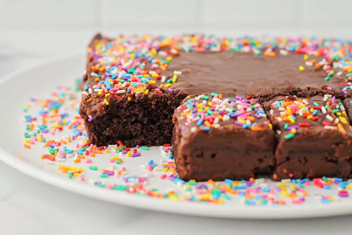 Close up of a one bowl chocolate cake baked and frosted with sprinkles on a platter with a piece removed.