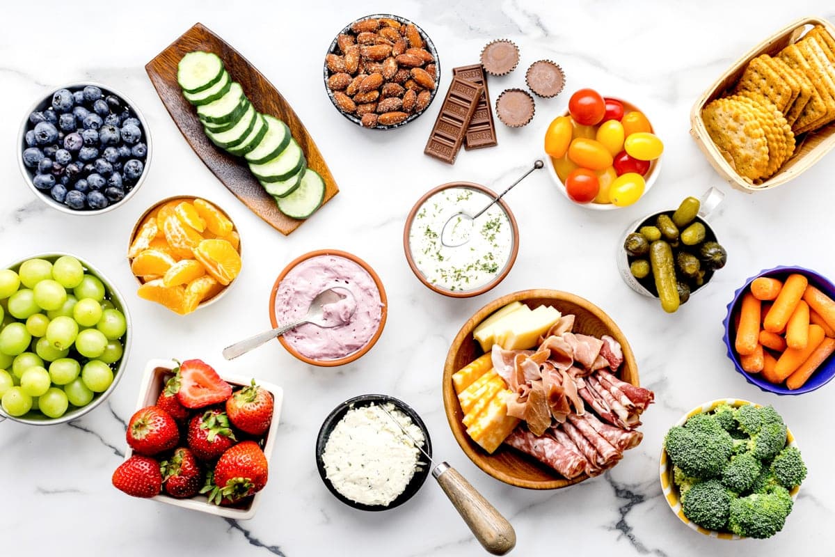 Charcuterie board ingredients set out in separate bowls, including blueberries, grapes, strawberries, cucumbers, carrots, broccoli, pickles, cherry tomatoes, almonds, crackers, chocolates, and dips. 