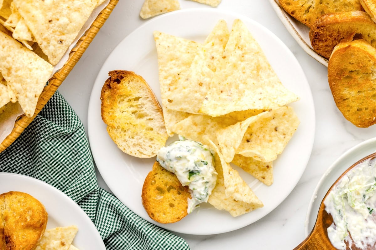 Bread and chips dipped in crockpot spinach artichoke dip on white plate.