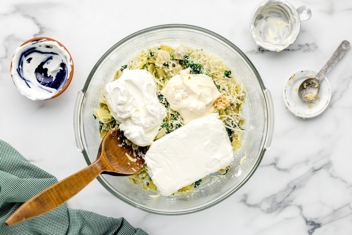 Wet ingredients added to spinach artichoke dip in glass bowl.