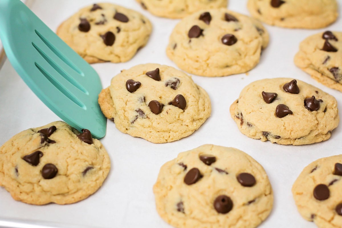 Chocolate chip cookies on baking sheet with spatula rounding out cookie edges.