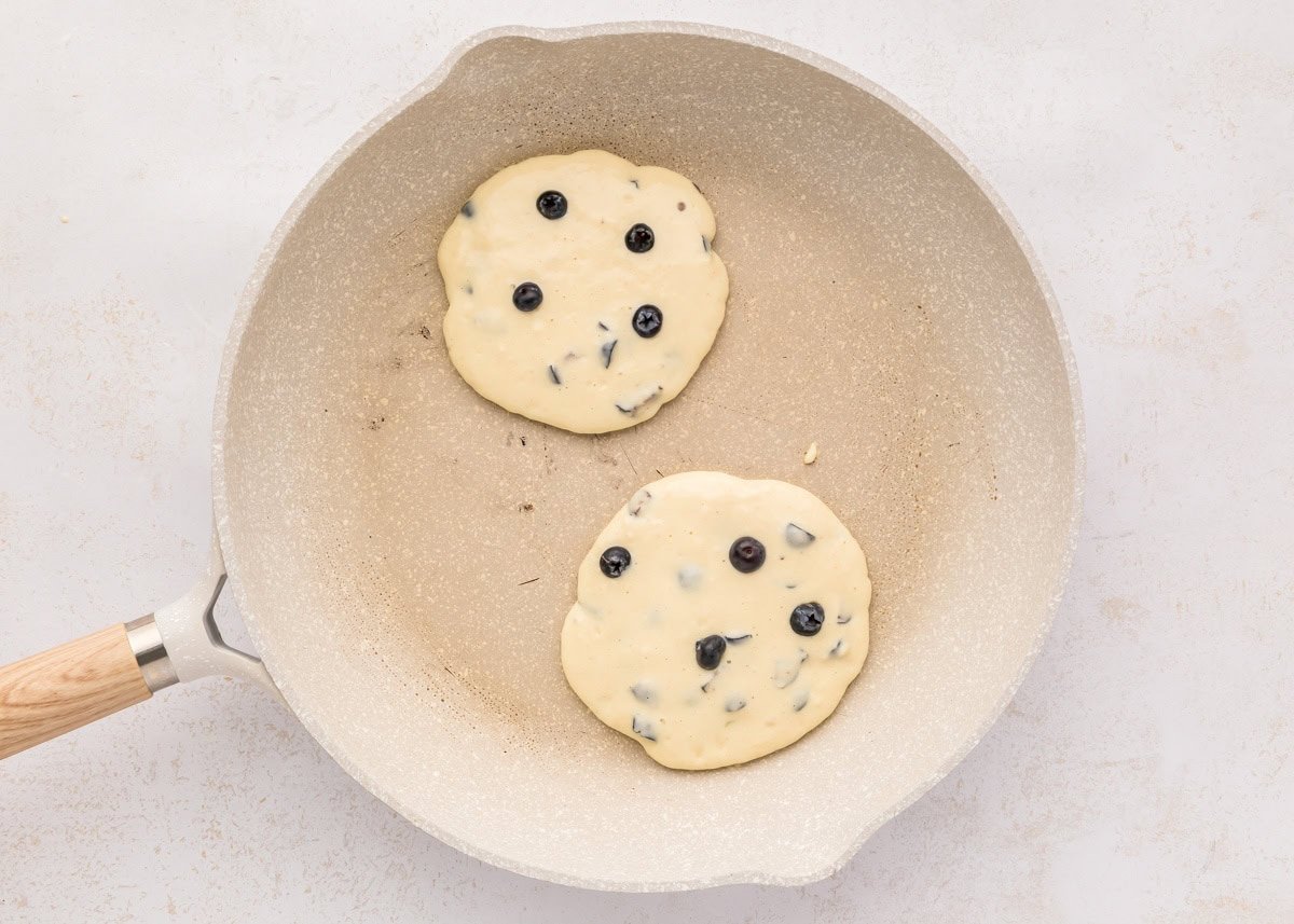 Two blueberry pancakes cooking in a skillet.