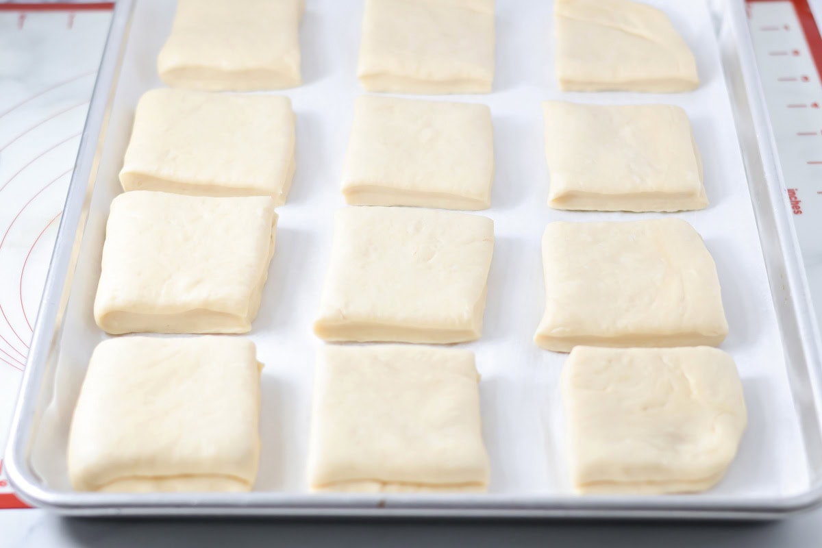 Uncooked squares of roll dough on a baking sheet.