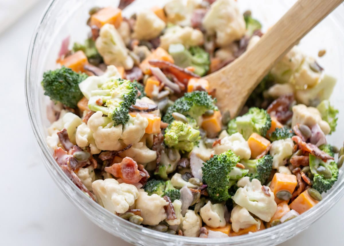 Broccoli cauliflower salad recipe ingredients in glass bowl with wooden spoon.