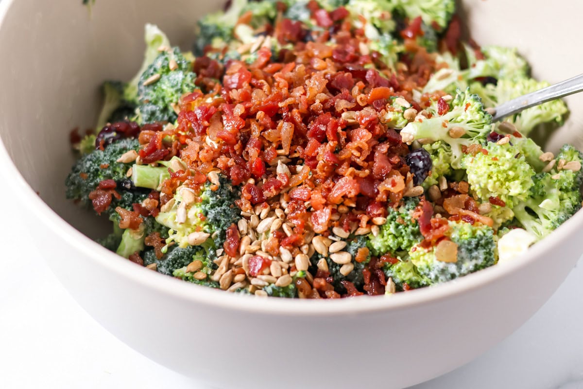 Adding sunflower seeds and bacon bits to the bowl of broccoli.