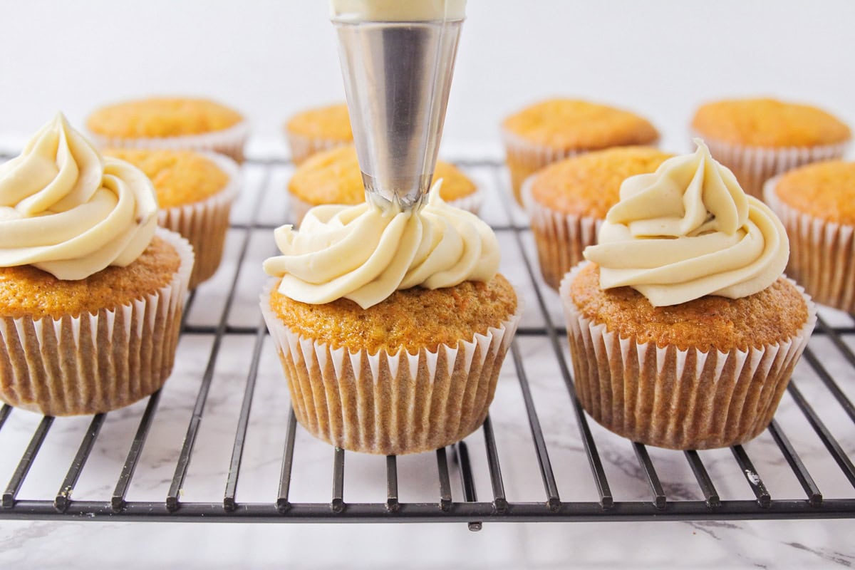 Frosting being piped on top cupcakes on wire rack.