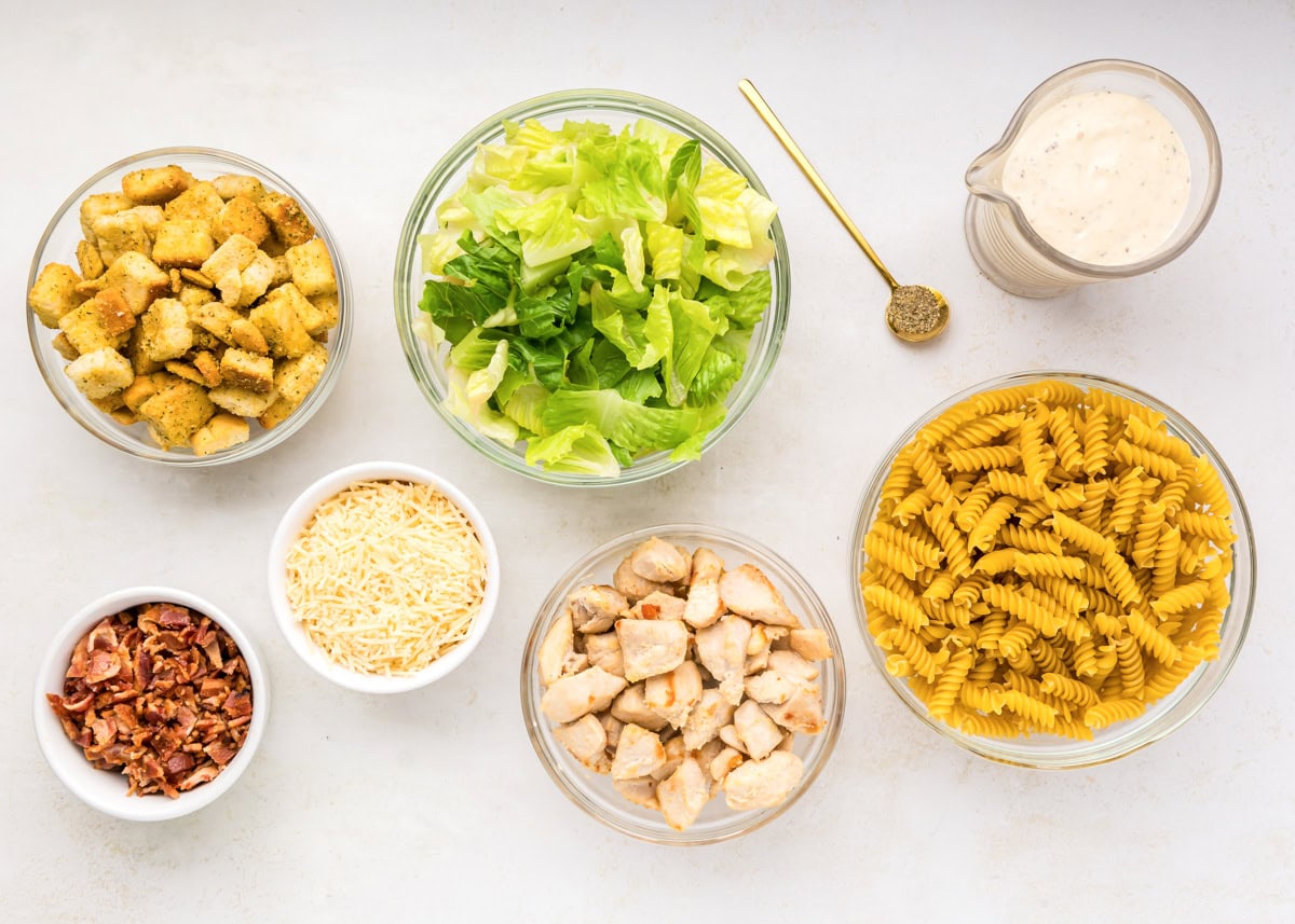 Lettuce, croutons, pasta, bacon, and dressing on a kitchen counter.