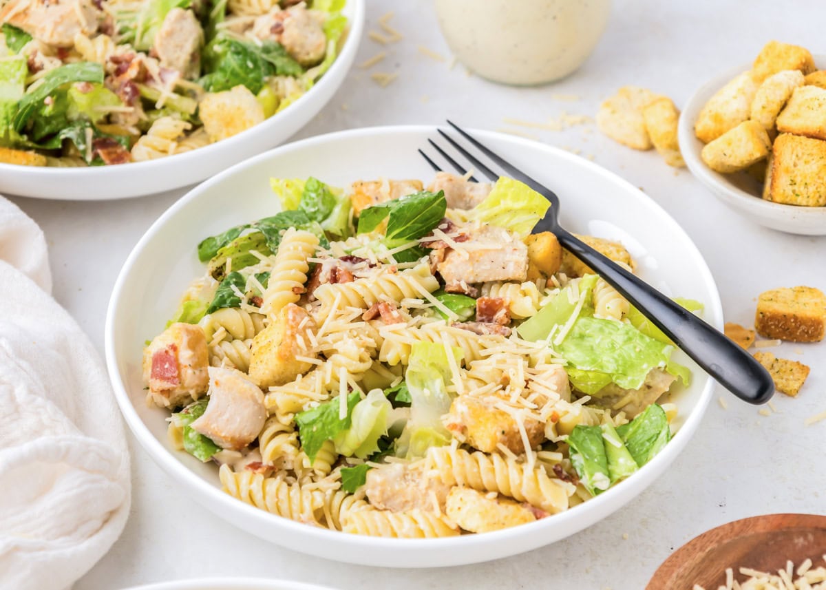 A serving of chicken Caesar pasta salad served in a white bowl with a black fork.