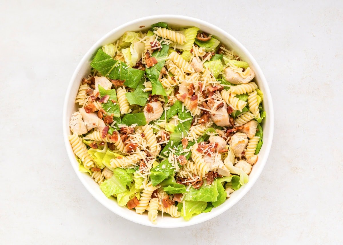 Combining lettuce, cooked pasta, cheese, and bacon in a white bowl.